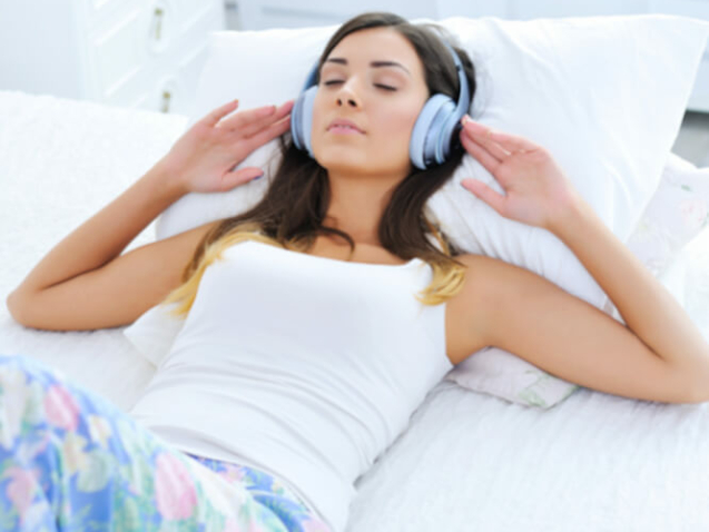 Eine junge dunkelhaarige Frau liegt entspannt auf dem Rücken und genießt mit geschlossenen Augen die Musik aus ihrem Kopfhörer.