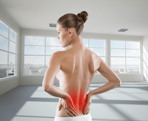 Eine junge blonde Frau steht mit dem Rücken zum Betrachter und hält sich mit beiden Händen den unteren Rücken. Ein großflächiger roter Punkt stellt die Schmerzen im unteren Rücken dar.
