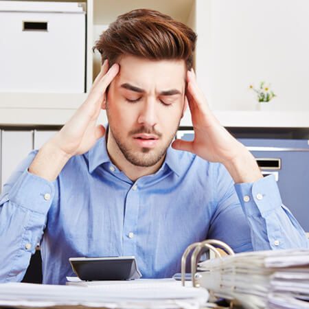 Junger Mann mit Rückenschmerzen durch sitzenden Lebensstil sitz am Schreibtisch und hält sich den Kopf.