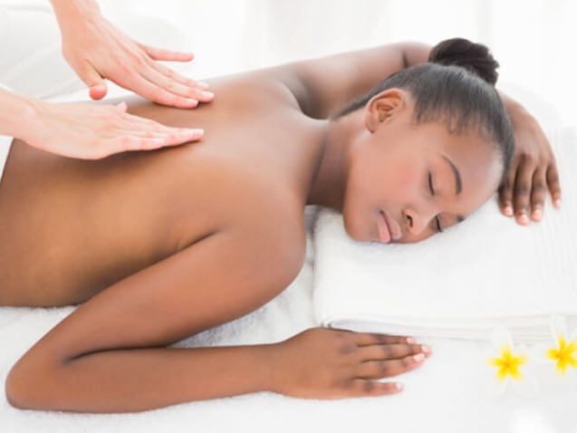 Eine junge dunkelhäutige Frau liegt entspannt auf einem Handtuch und bekommt eine Massage am Rücken.