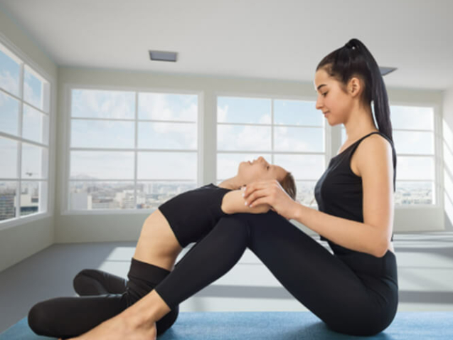 Eine junge orientalisch aussehende Frau sitzt nach links gewendet am Boden. Sie hält ein Mädchen an den Armen und streckt dessen Rücken über Ihre Knie.