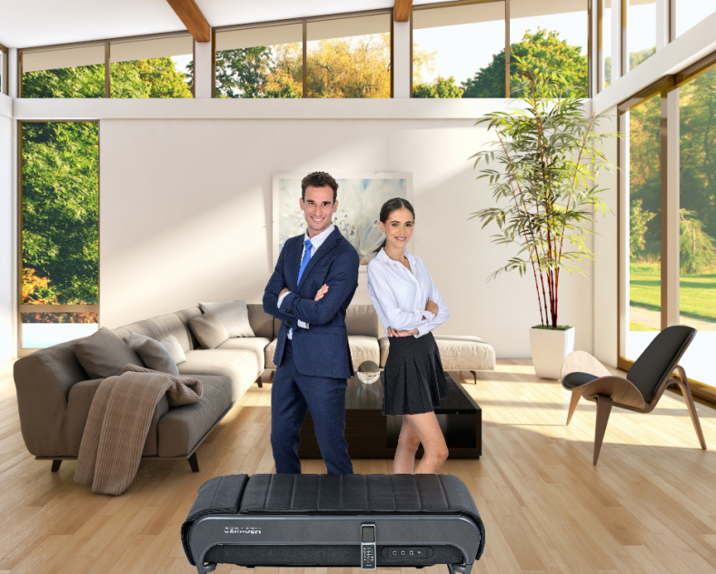 CERAGEM-Anwendungsraum: Wohnzimmer. Ein lichtdurchfluteter Raum mit Sofa, kleinem Tisch und Stuhl. Im Hintergrund sind ein Bild und eine große Pflanze. Ein Mann und eine Frau stehen lächelnd hinter der CERAGEM V4 Liege.